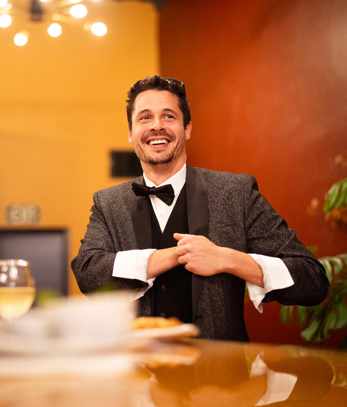 The Comparable Jason Sims A smiling man with dark hair and facial hair wearing a sparkly tux and black bowtie stands behind a counter with his hand inside his coat pocket.